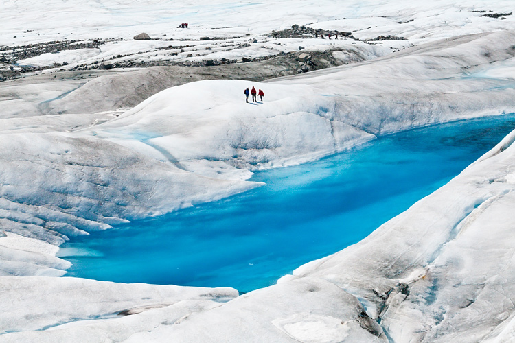 Figures walking by a bright blue melt pool on a glacier in Juneau